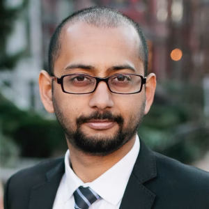 A man with a short beard and glasses is wearing a black suit and a white shirt with a tie. He is standing outdoors, with blurred greenery and buildings in the background